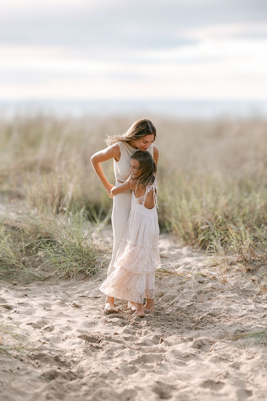 A mother in a gray dress hugs her young daughter in a white dress on a sandy beach path surrounded by dune grass.