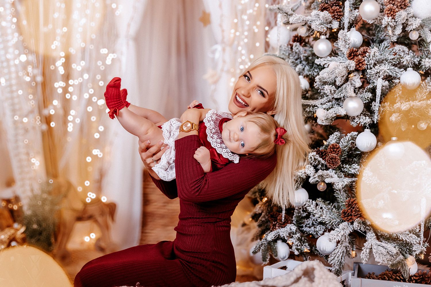 Smiling blonde mother holding baby girl in matching red outfits near decorated Christmas tree with bokeh lights.