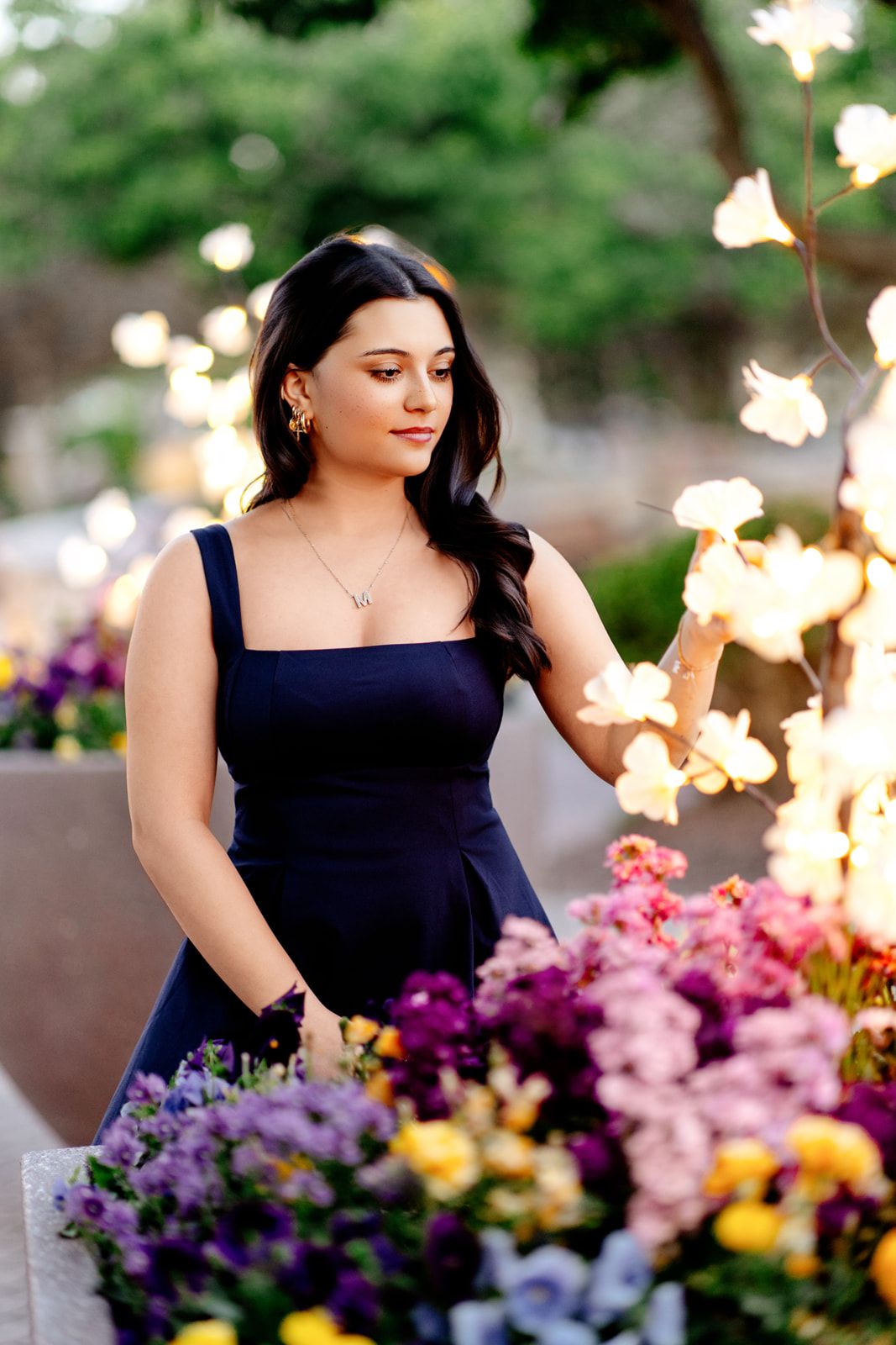 Young woman in navy dress admiring colorful flowers surrounded by soft bokeh lights.