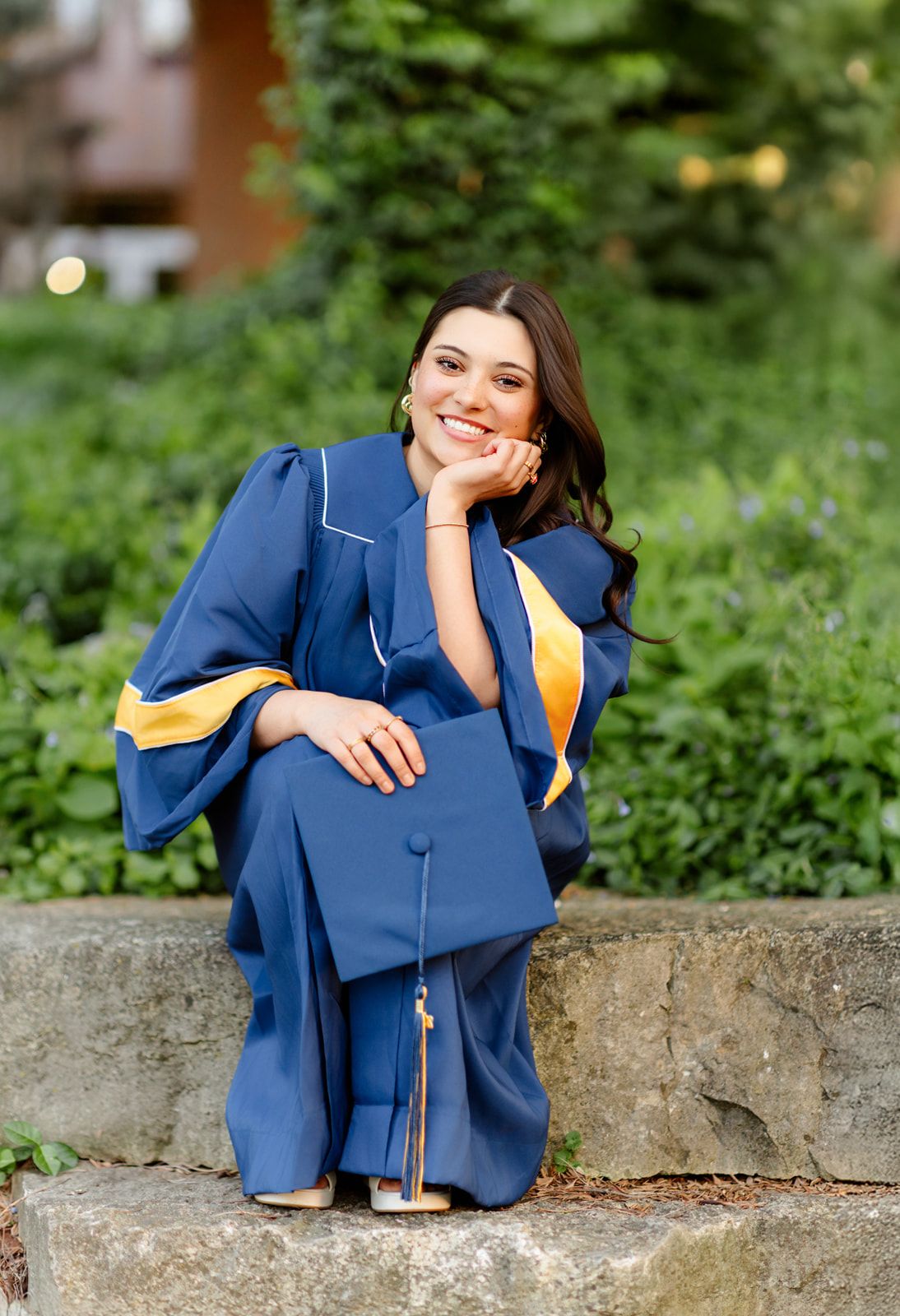 Happy graduate in blue and gold gown sitting on stone wall surrounded by lush greenery.