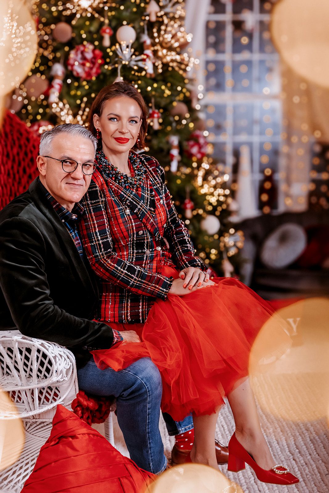 Couple posing together in front of decorated Christmas tree, woman in red plaid outfit and tulle skirt, man in dark velvet blazer.