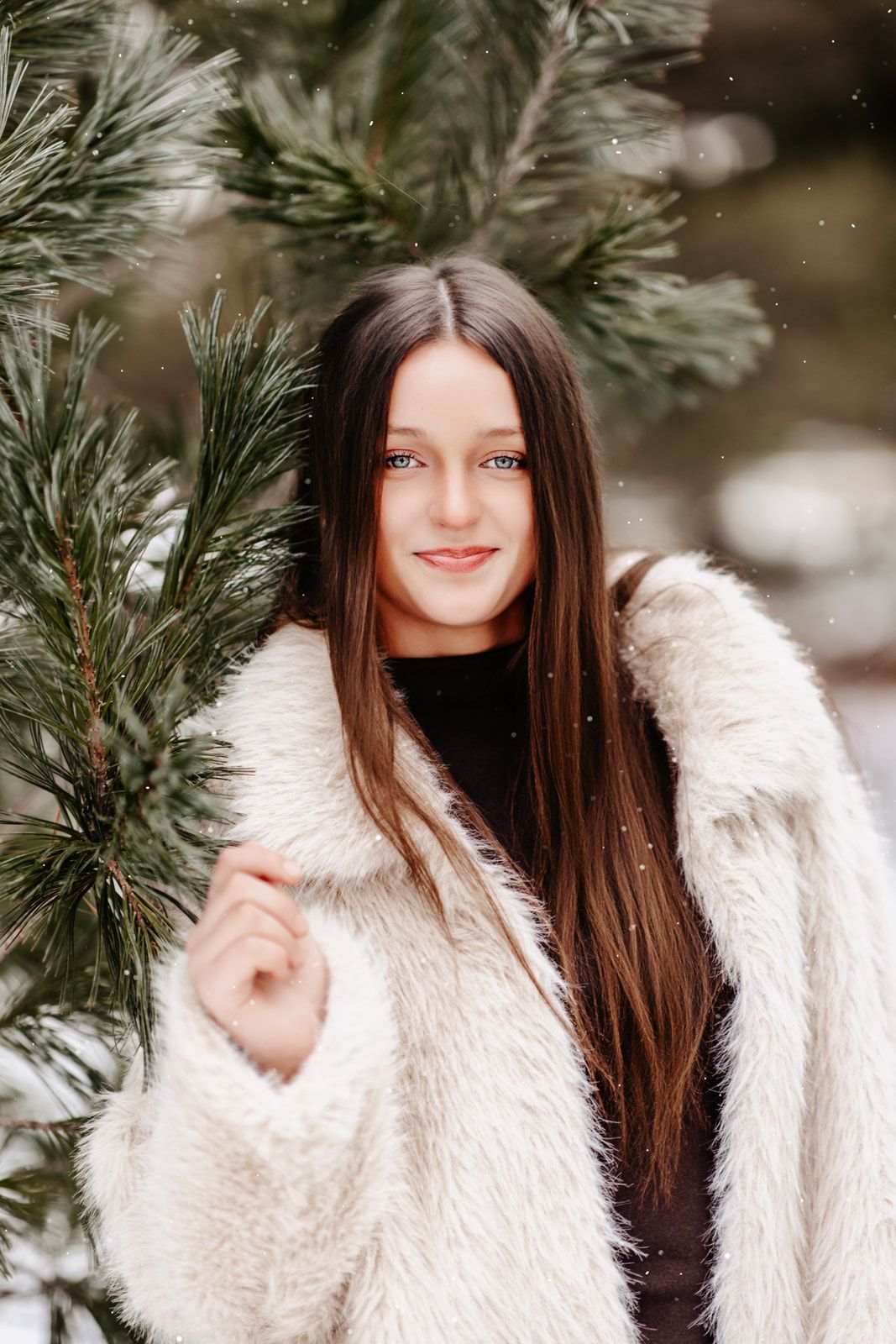 Young woman with long dark hair smiling in white fluffy coat among snow-dusted pine branches in winter forest.