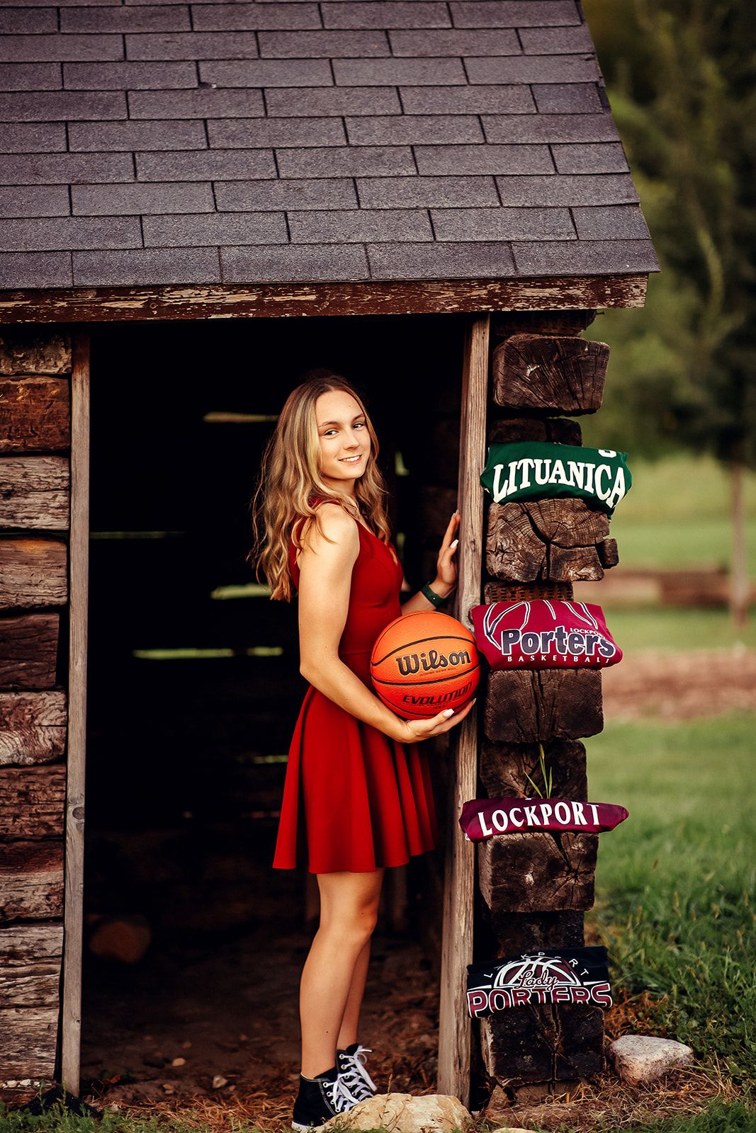 Smiling teen in red dress holding Wilson basketball beside team signs at rustic log shed.