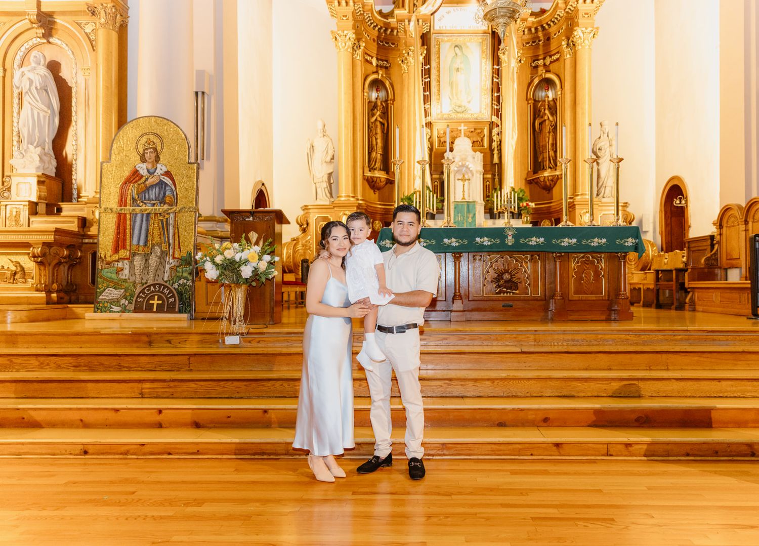 Couple in white outfits holds young child inside elegant Catholic church with golden altar.