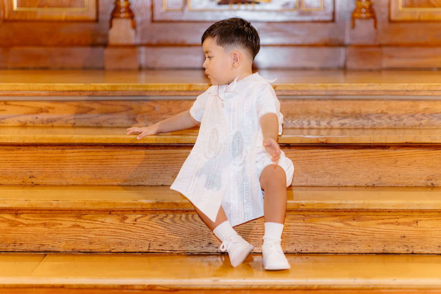 Toddler in white baptism outfit sitting on wooden church steps looking to the side.