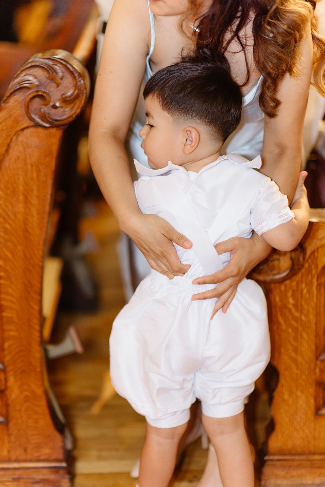 Young boy in white baptism outfit standing between church pews, held by a woman in a blue dress.