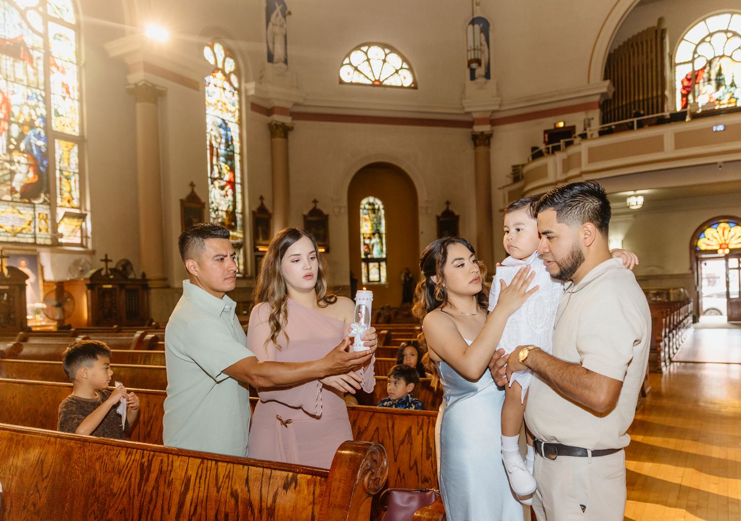 Family gathered in a beautiful church for a baptism ceremony, holding a candle near wooden pews with stained glass windows.