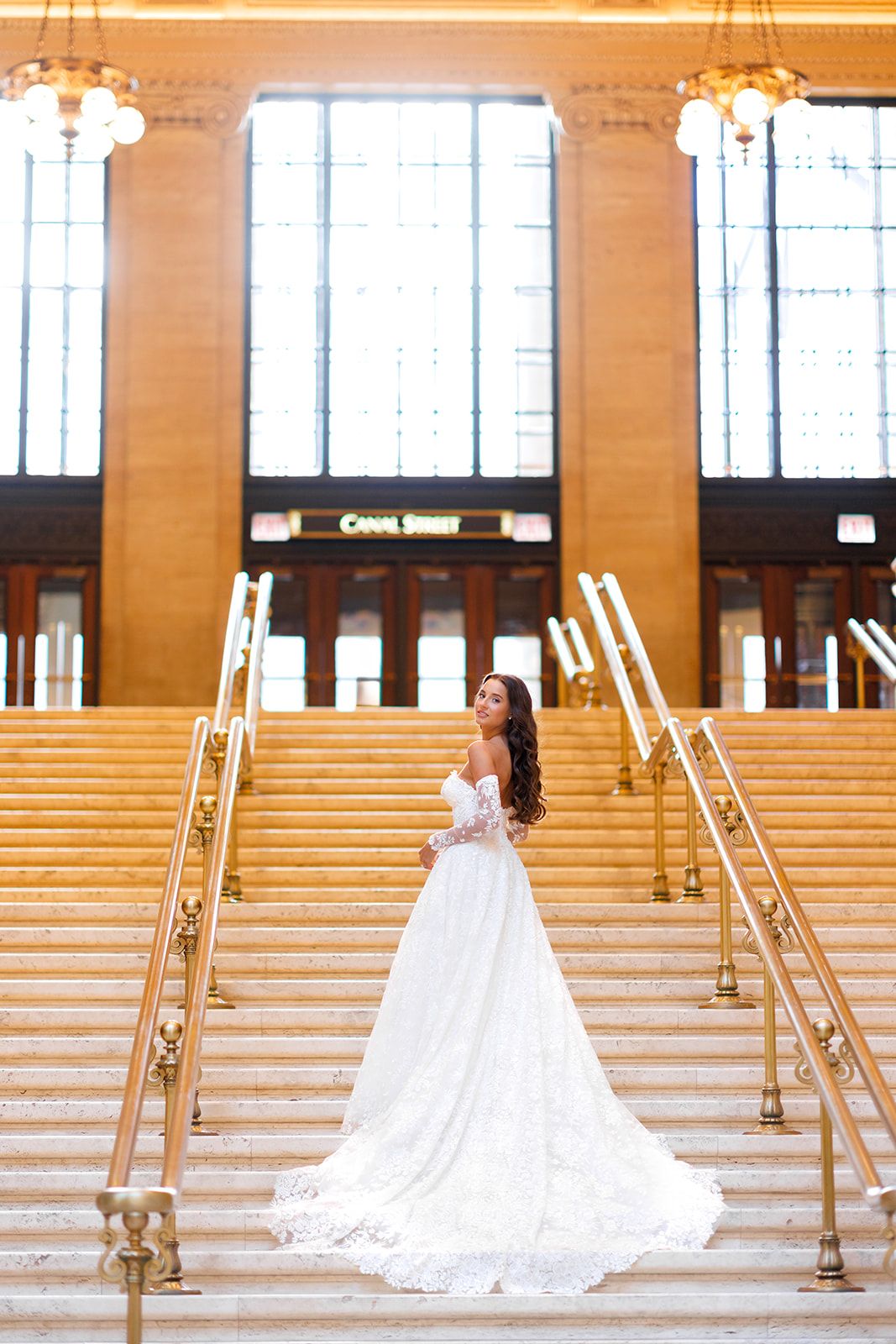 Bride in white lace gown stands on grand staircase at Canal Street station, chandeliers glowing above.