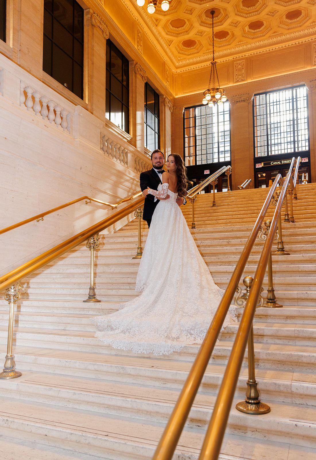 Bride and groom pose on grand marble staircase with gold railings in elegant wedding venue.