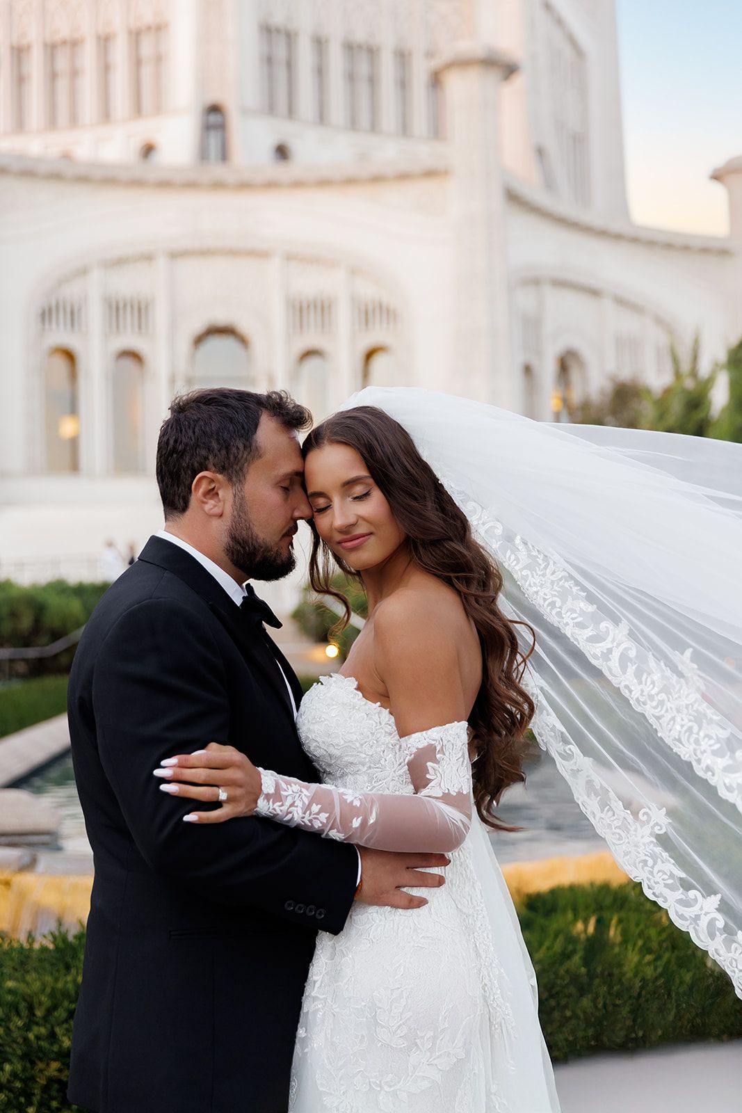 Bride and groom share a romantic forehead touch in front of a grand white building, bride wearing lace gown and veil.