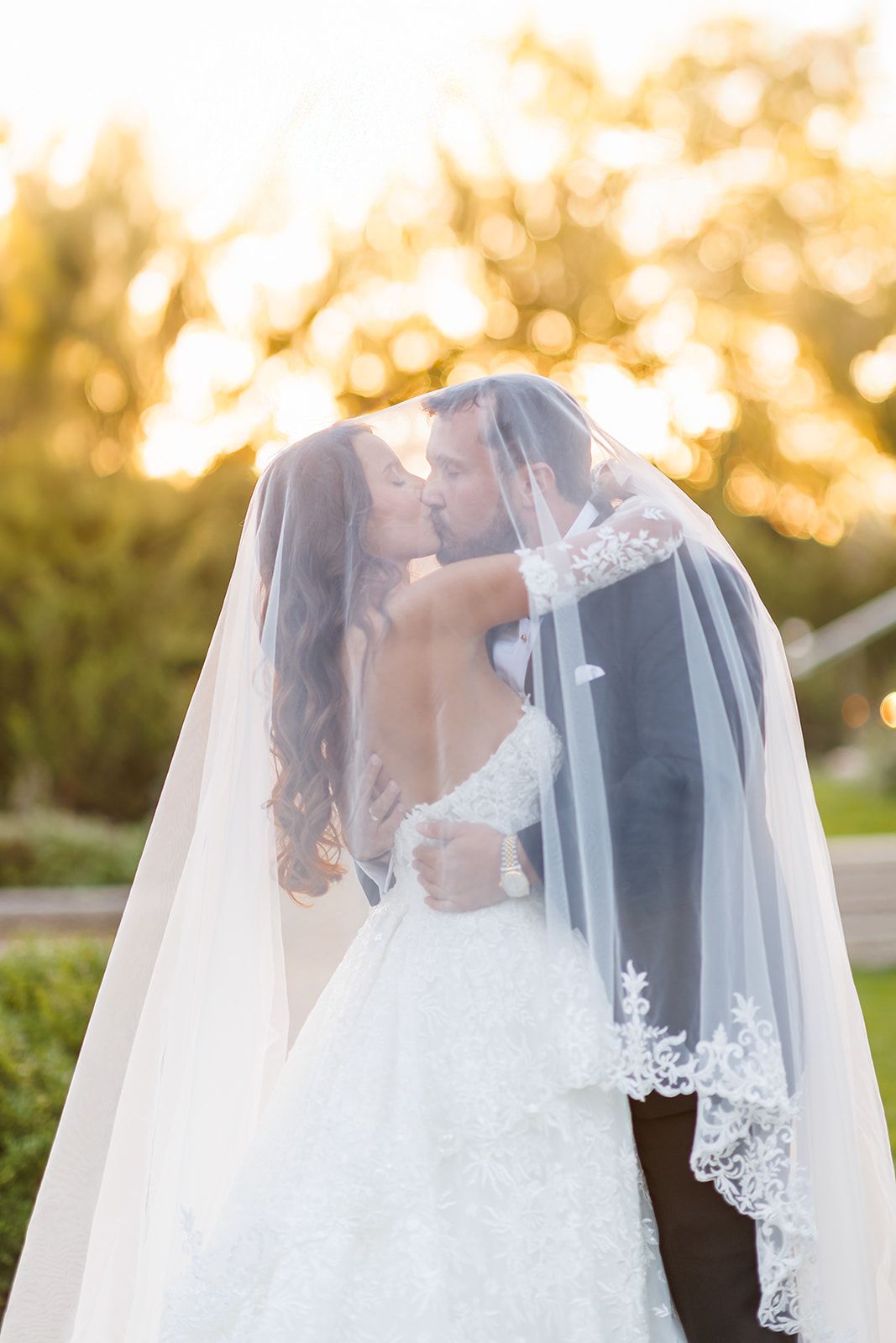 Newlyweds embrace and kiss beneath a flowing bridal veil during a stunning golden sunset.