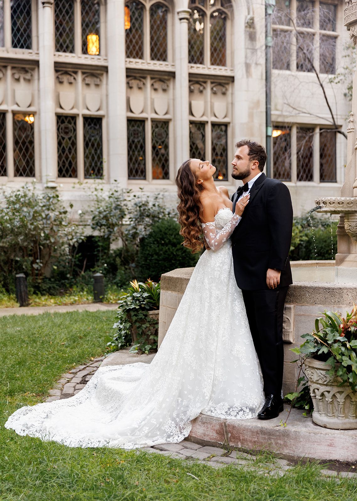 Bride in off-shoulder lace wedding gown gazes up at groom in tuxedo in gothic castle garden setting.