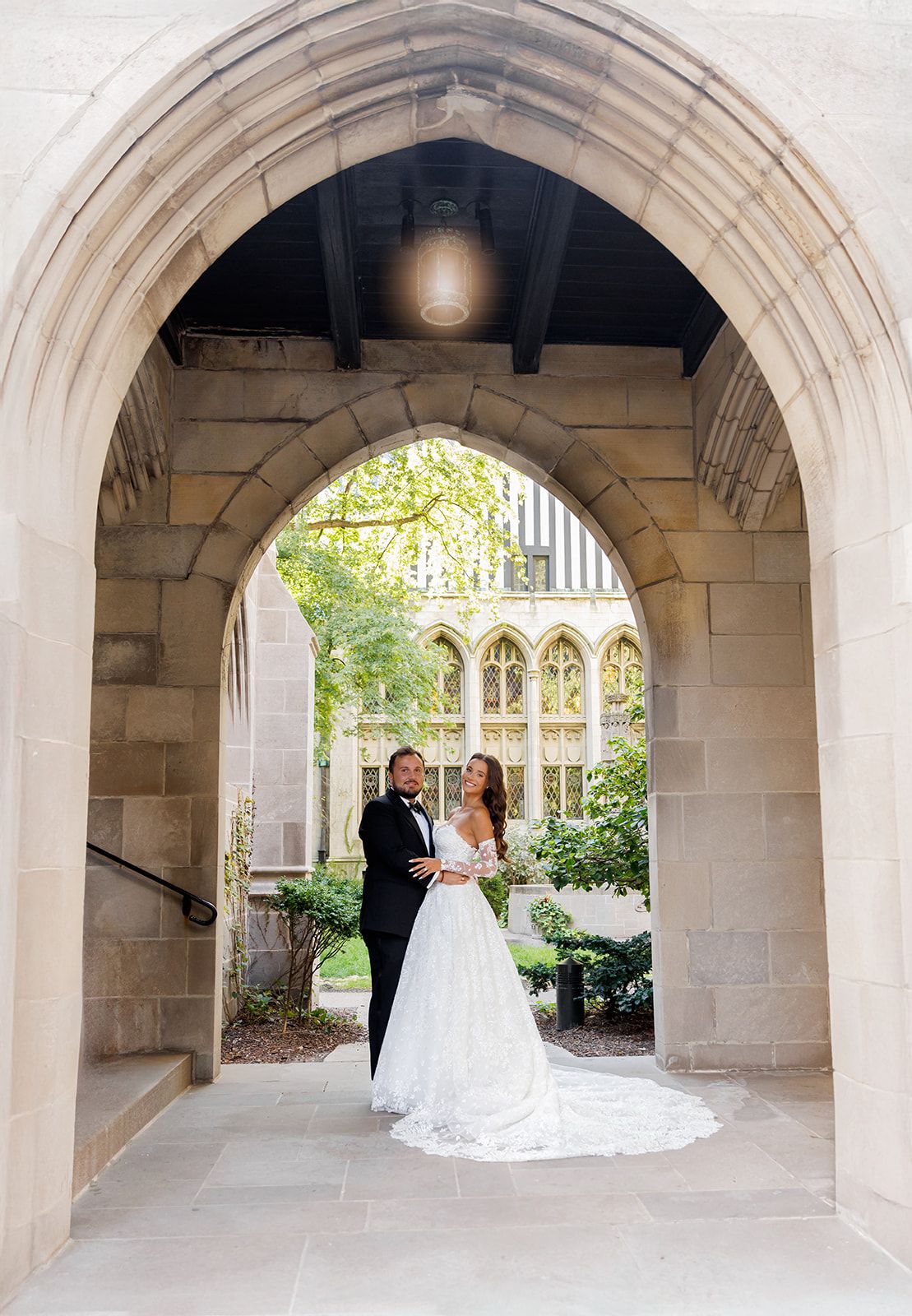 Bride and groom embrace under a grand Gothic stone arch framing a lush courtyard garden behind them.