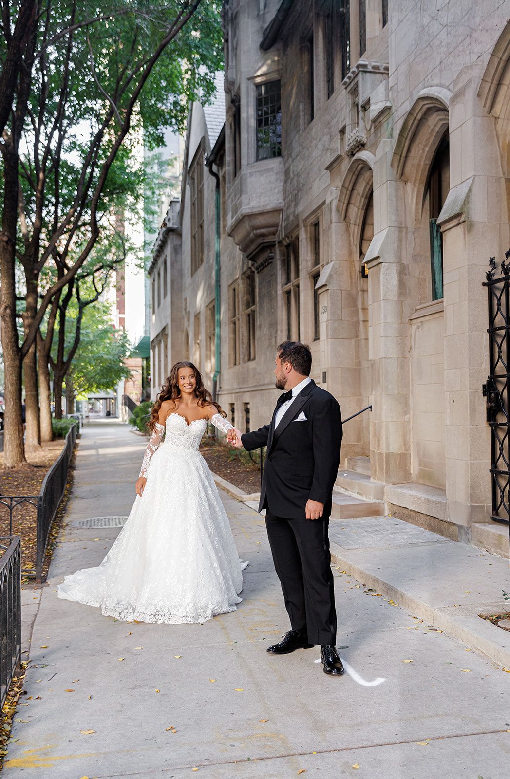 Bride in white lace ball gown holds groom's hand in black tuxedo on a tree-lined sidewalk beside a stone building.
