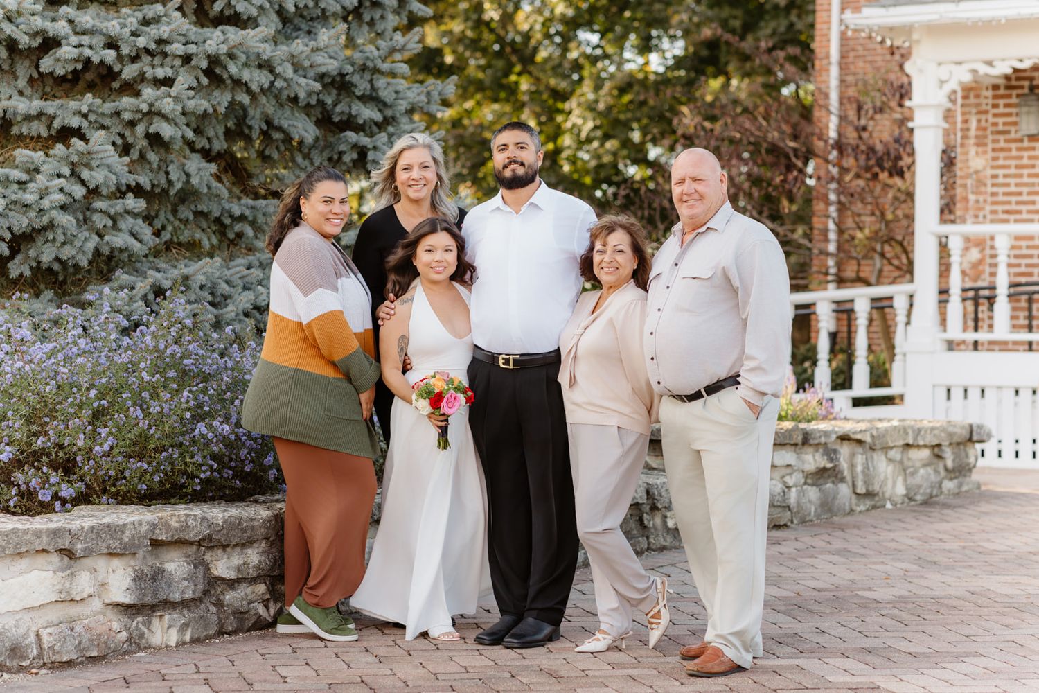 Wedding party of five poses outdoors, bride in white holding bouquet with groom and family near gazebo.