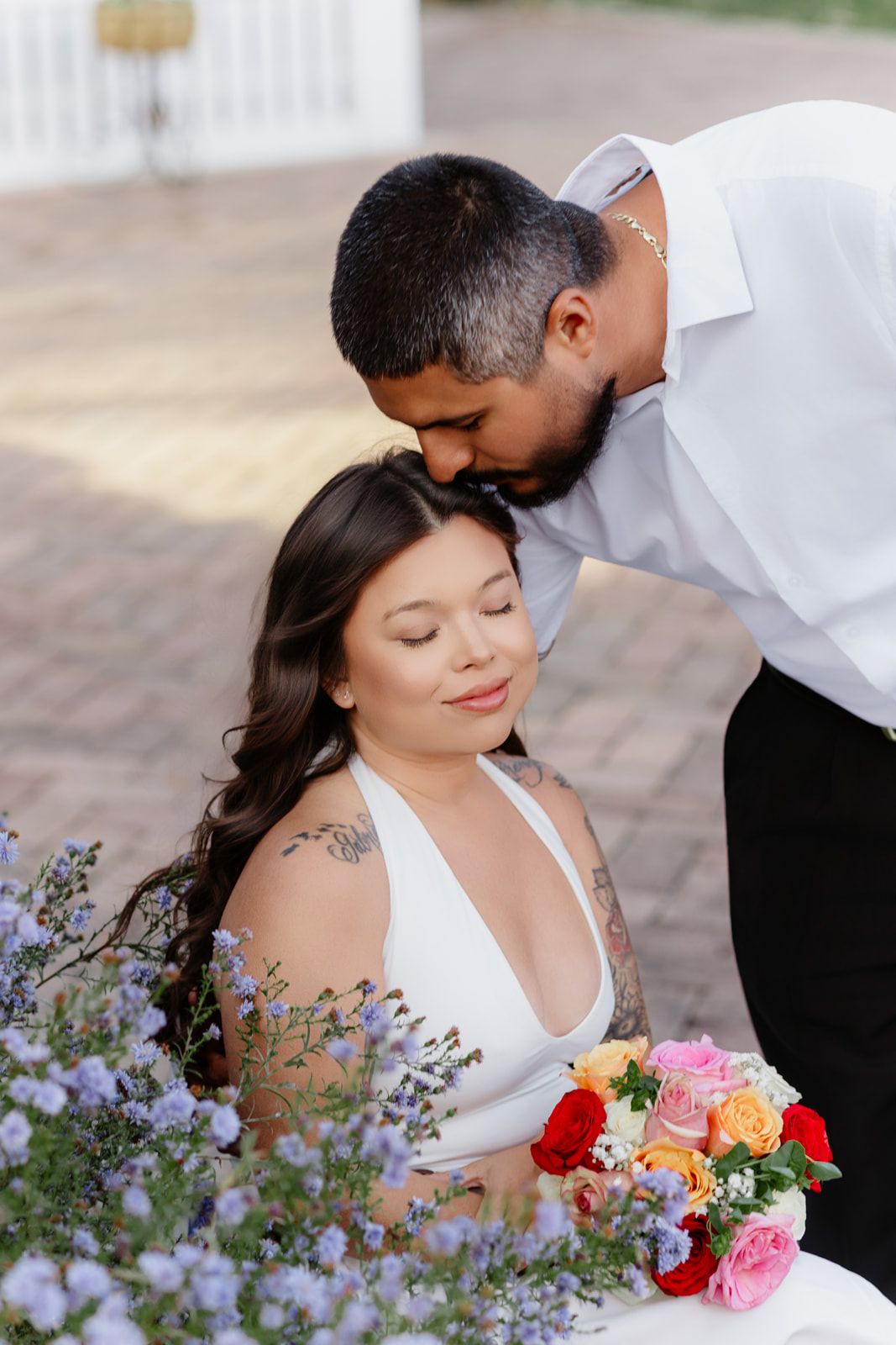 Groom kissing bride on head while she holds colorful bouquet of roses outdoors at wedding ceremony.