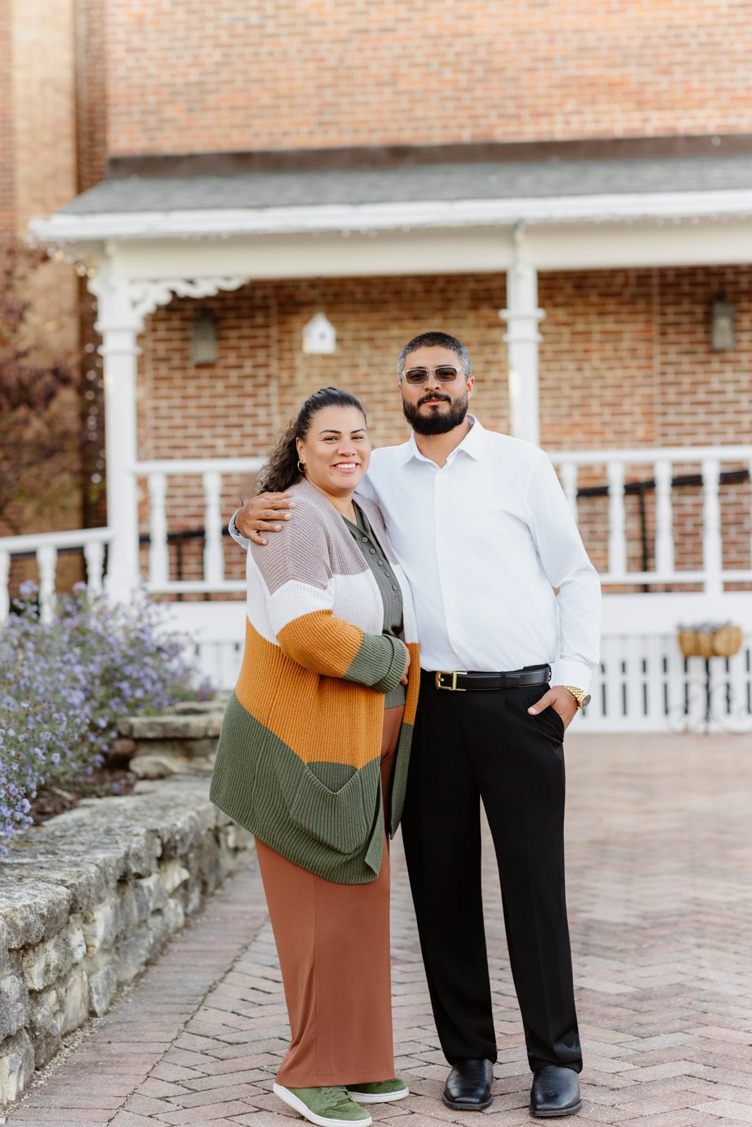 Couple poses together in front of brick home, woman in colorful cardigan and man in white shirt smiling warmly.