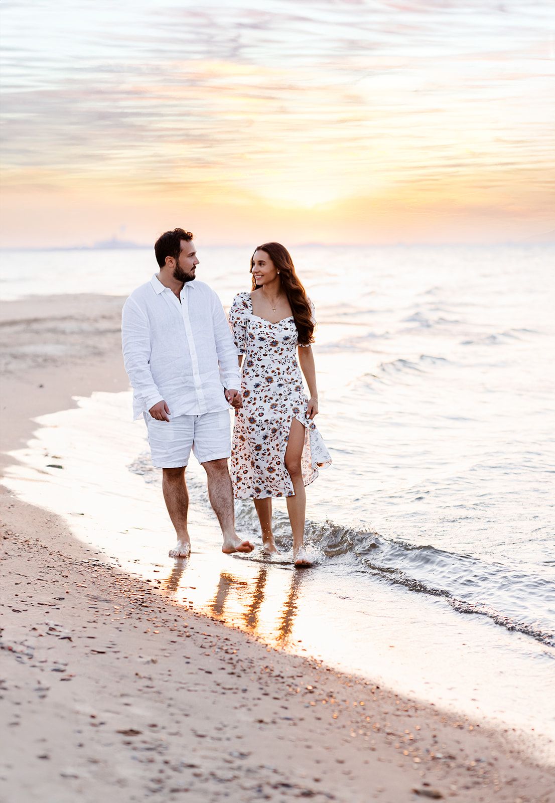 Happy couple in white outfits walking hand in hand along shoreline at colorful sunset.