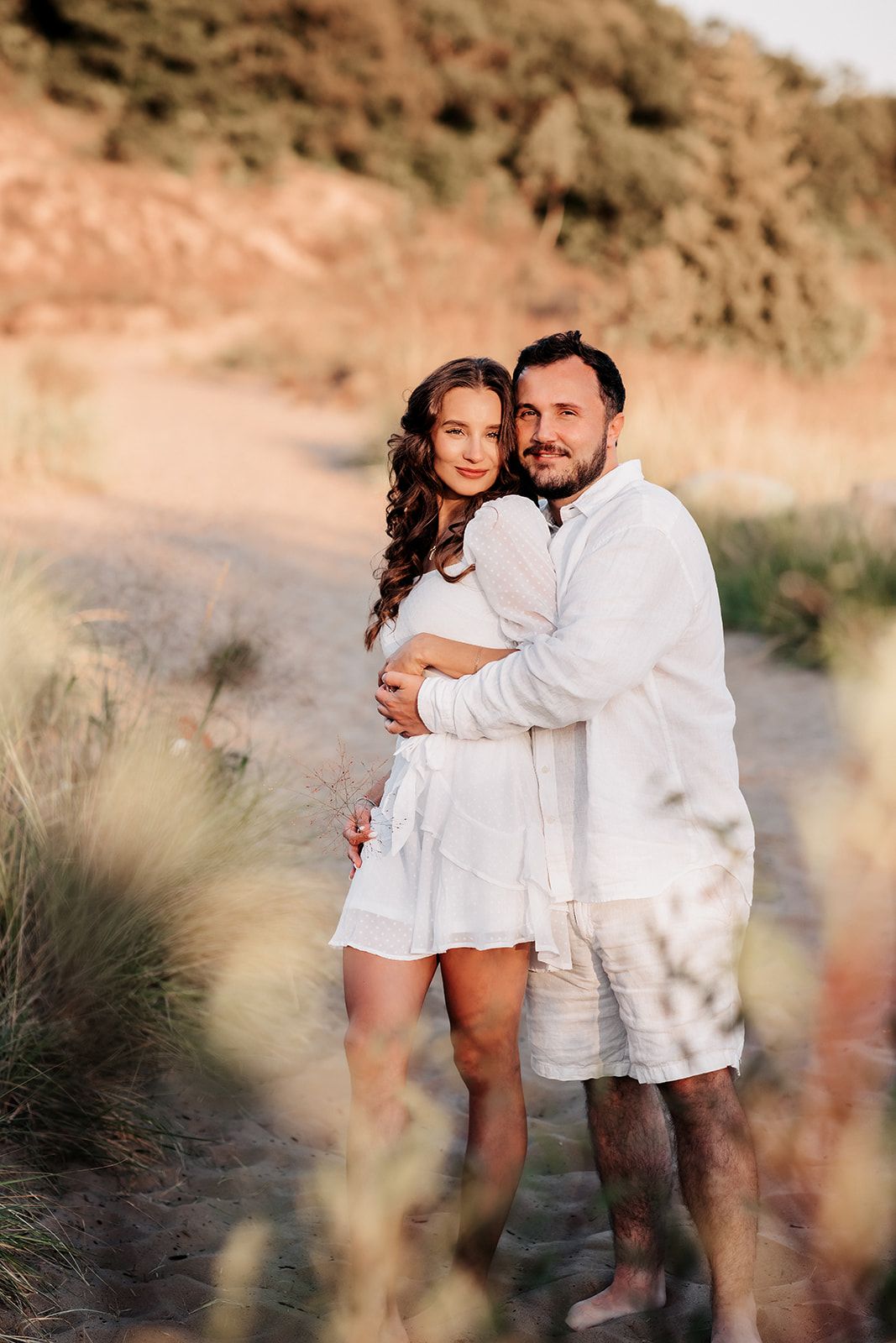 Couple in white outfits embracing on a sandy beach path with golden grass and dunes in the background.