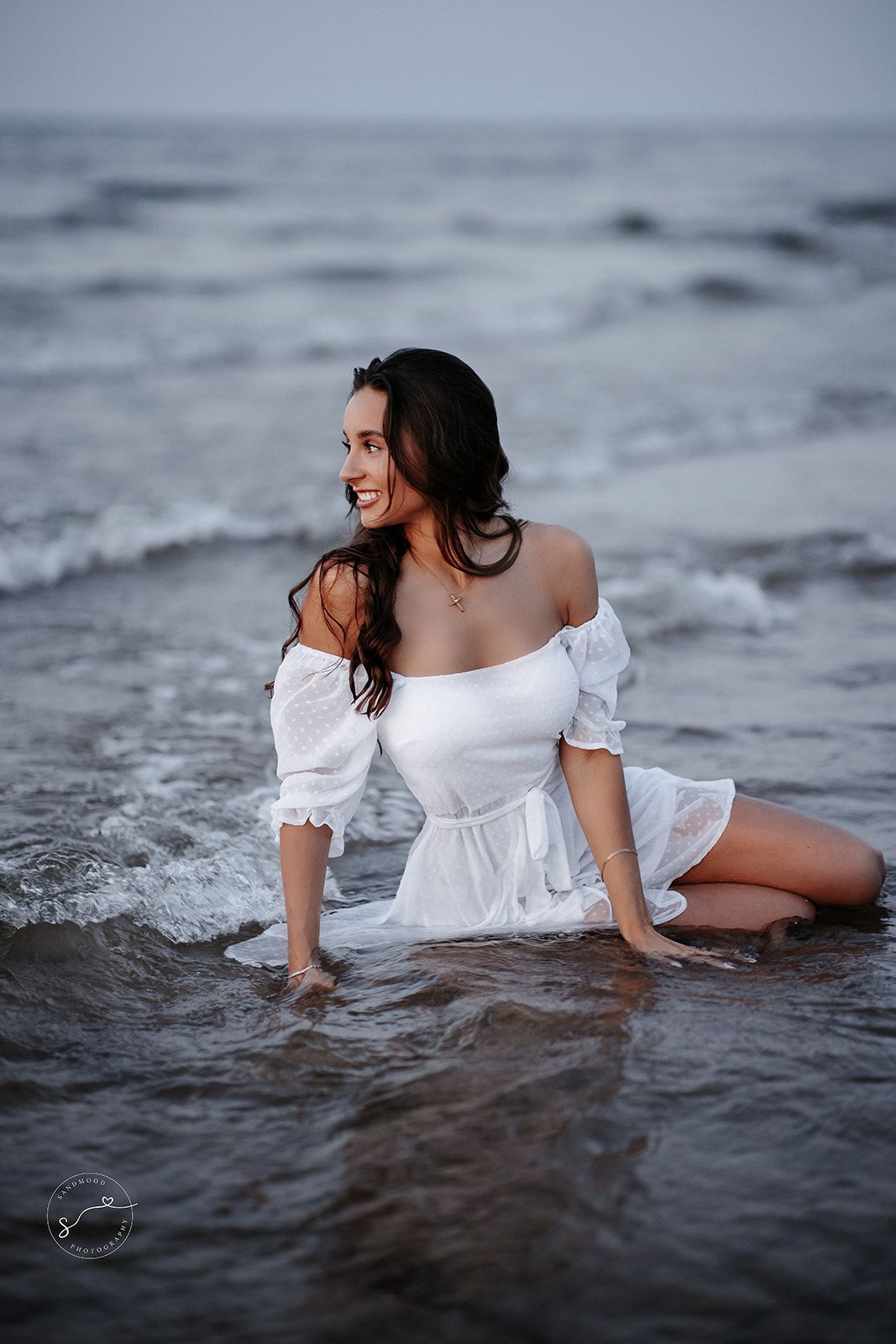 A smiling woman in a white off-shoulder dress sits gracefully in ocean waves on a beach.