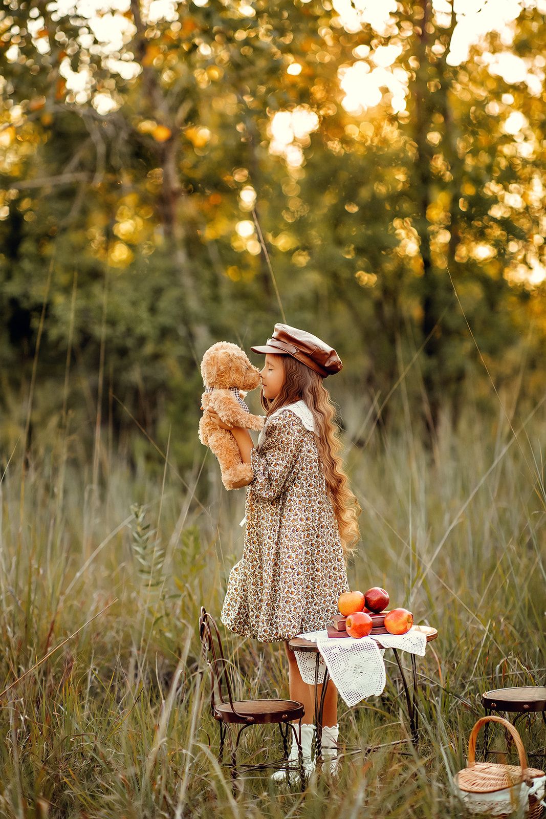 A little girl in a floral dress and hat holds a teddy bear at an outdoor picnic table surrounded by autumn forest.