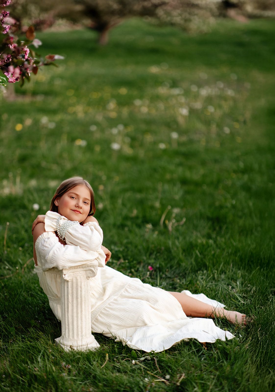 Young girl in white dress sitting on grass leaning against a pillar, surrounded by blooming spring trees.