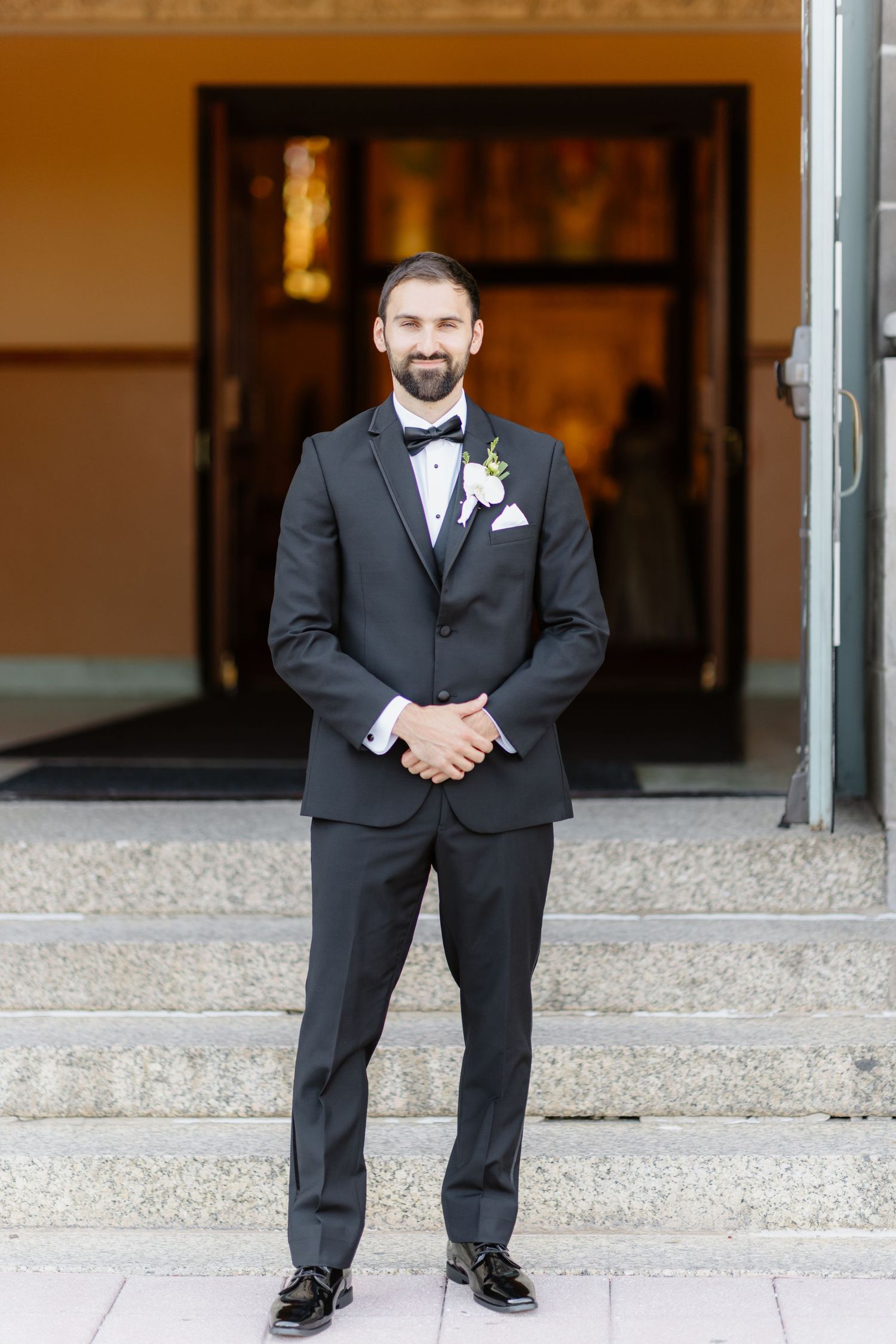 Smiling bearded groom in black tuxedo with boutonniere posing on church steps before wedding.
