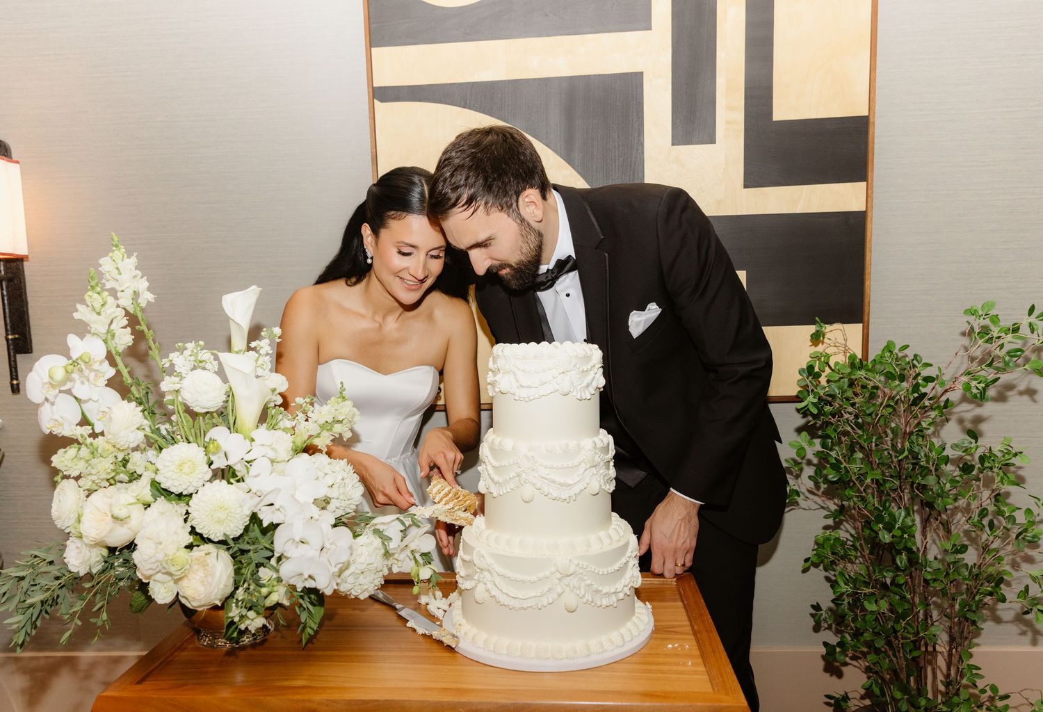 Bride and groom cutting their beautiful white tiered wedding cake surrounded by white floral arrangements.