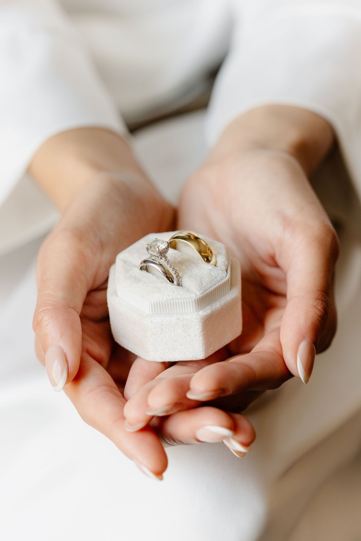 Hands holding a white velvet ring box with a diamond engagement ring and gold wedding band.