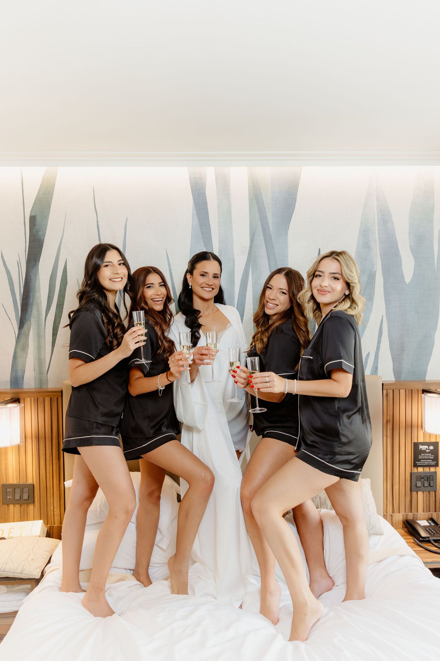 Bride in white robe celebrates with four bridesmaids in matching black satin pajamas, toasting champagne on hotel bed.