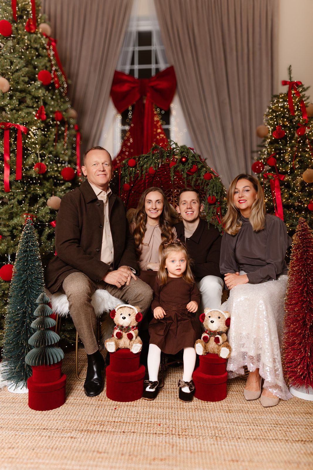 Family of five posing for a Christmas photo surrounded by decorated trees with red bows and ornaments, with teddy bears and a toddler girl.