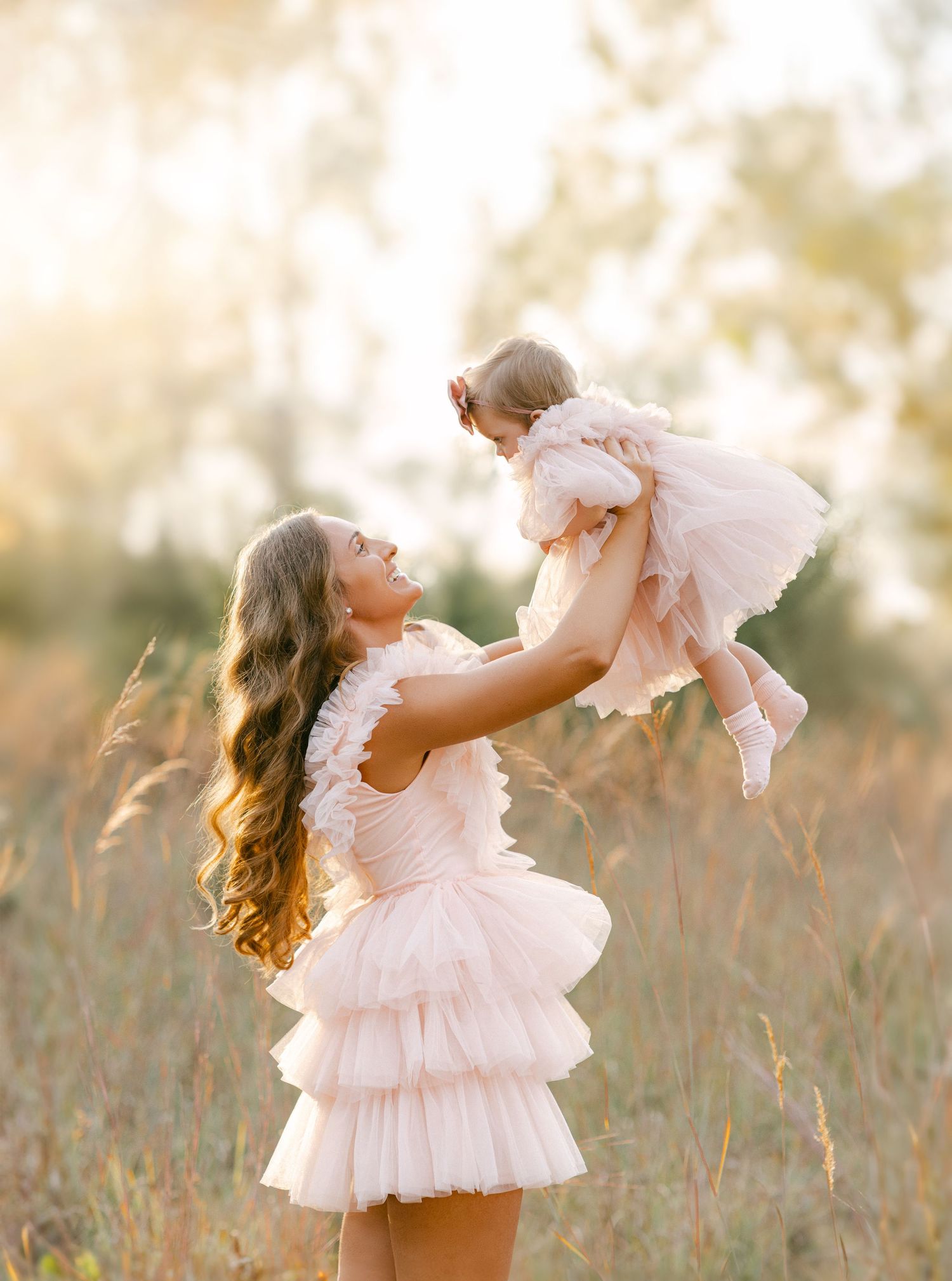 A mother in a pink ruffle dress lifts her baby girl in a matching dress outdoors in a golden sunlit field.