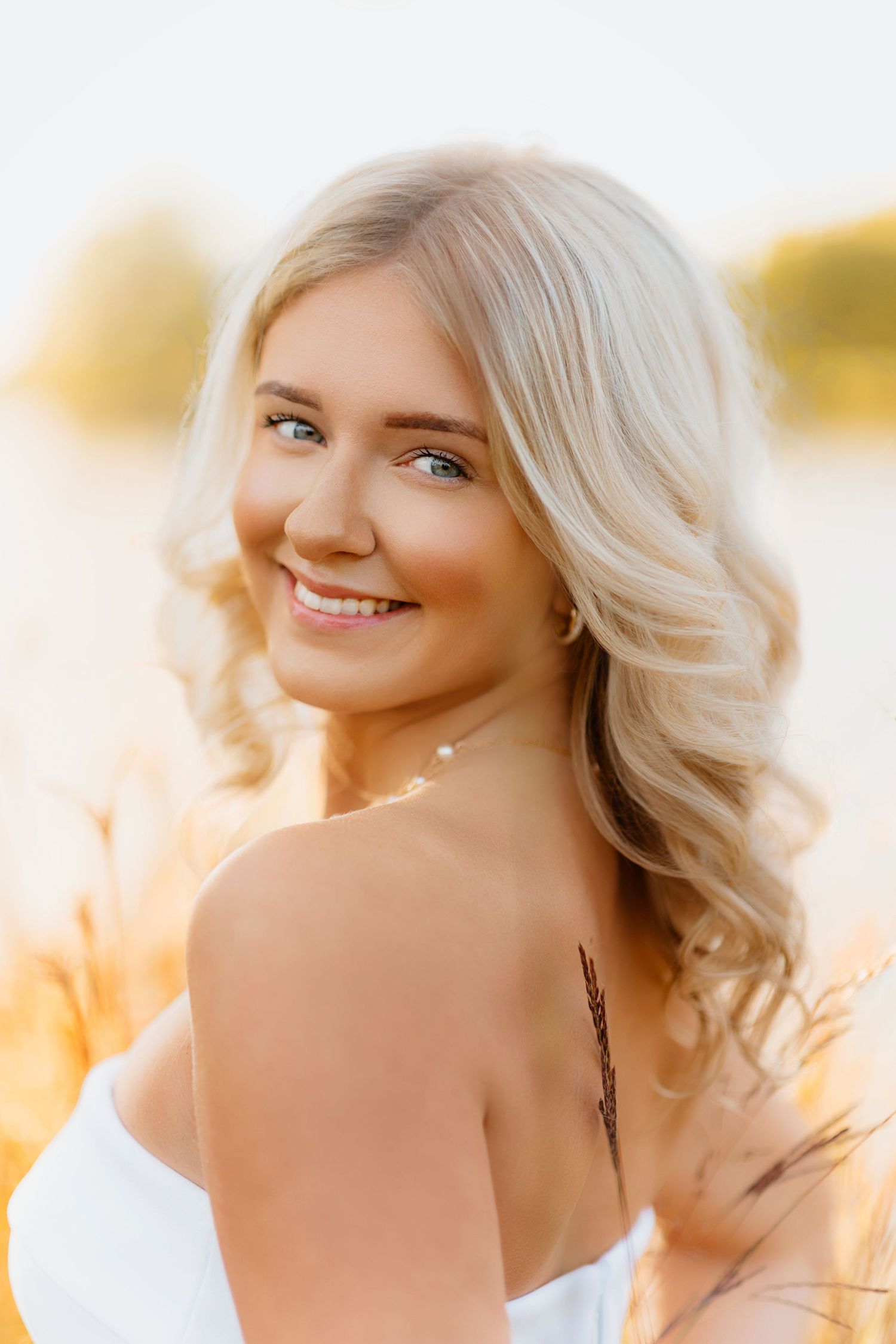 Smiling blonde woman in white strapless dress in golden sunlit grass field at sunset.