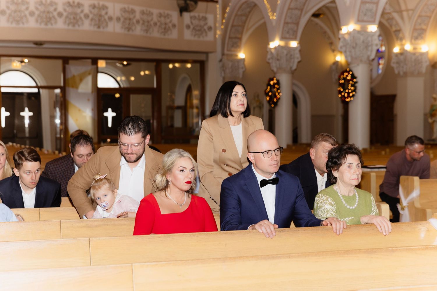 Family members seated in church pews during wedding ceremony with woman in red dress foreground.
