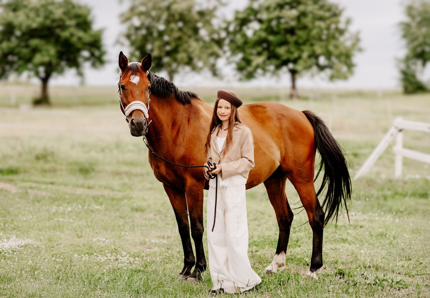 Woman in beret and cream outfit stands with a brown horse in a lush green field with trees.