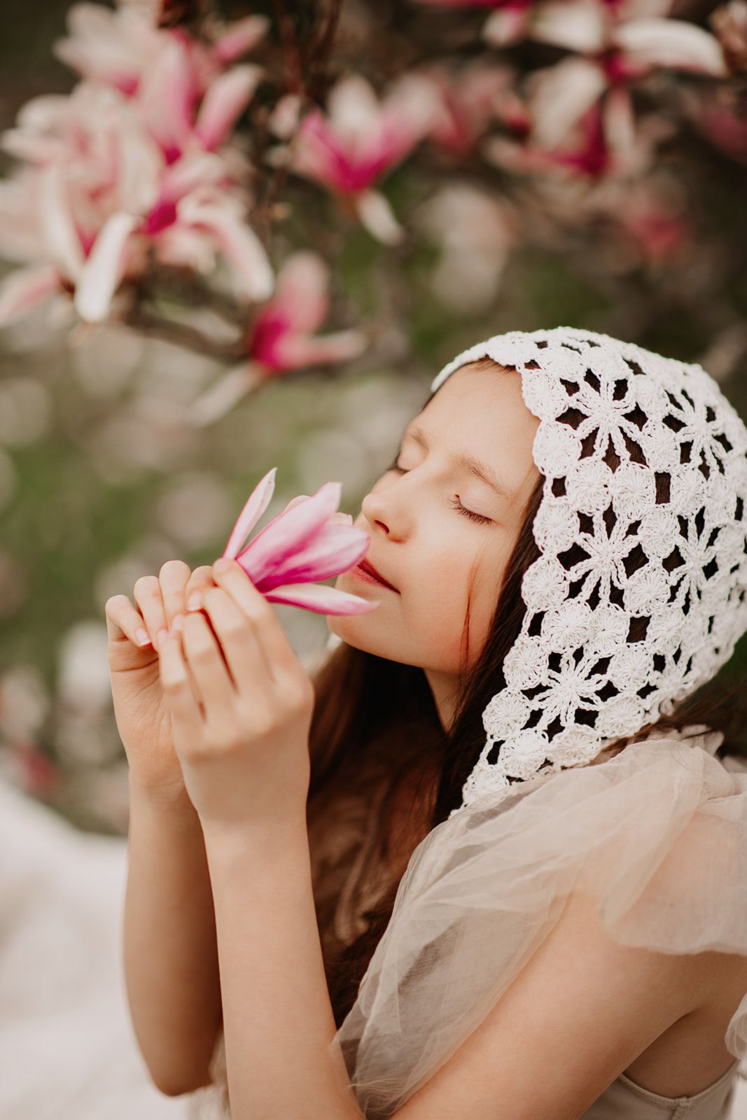 A young girl with closed eyes smells a pink magnolia flower, wearing a white crochet bonnet among blooming trees.