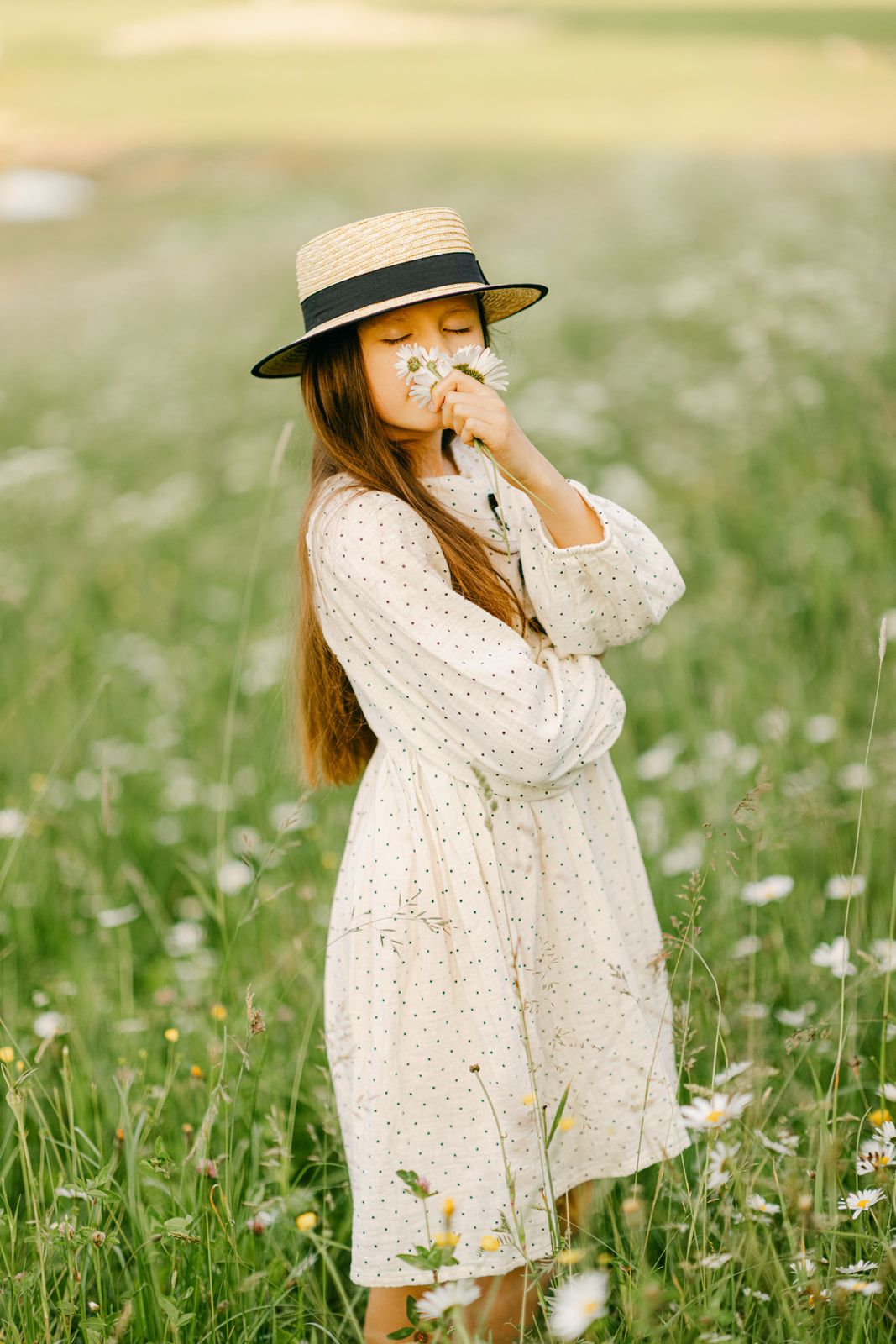 Girl in white polka dot dress and straw hat smells wildflowers while standing in daisy field.