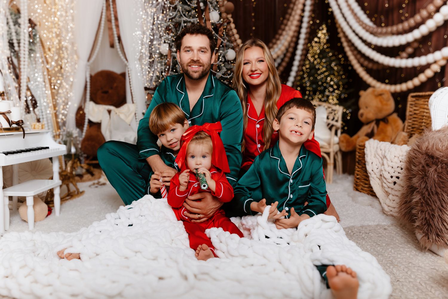 Family of five in matching green and red Christmas pajamas sitting together by a decorated holiday tree.