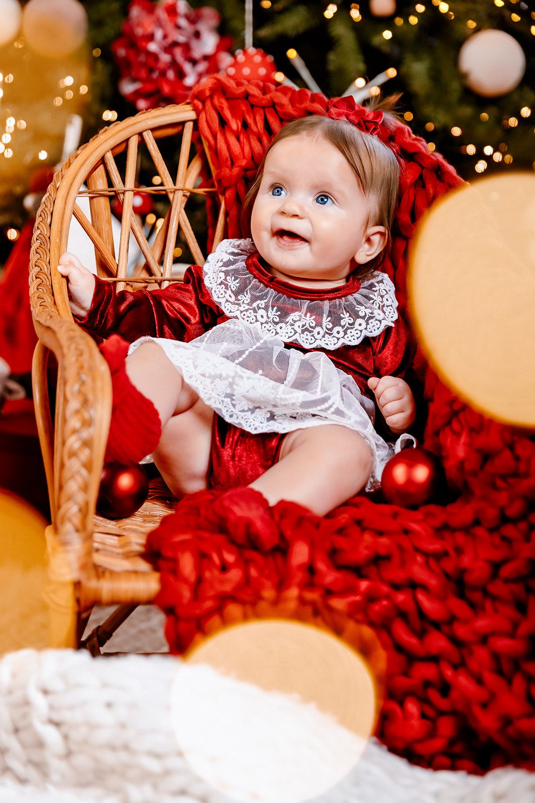 Adorable baby girl in red velvet dress with lace collar sitting in wicker chair by decorated Christmas tree.