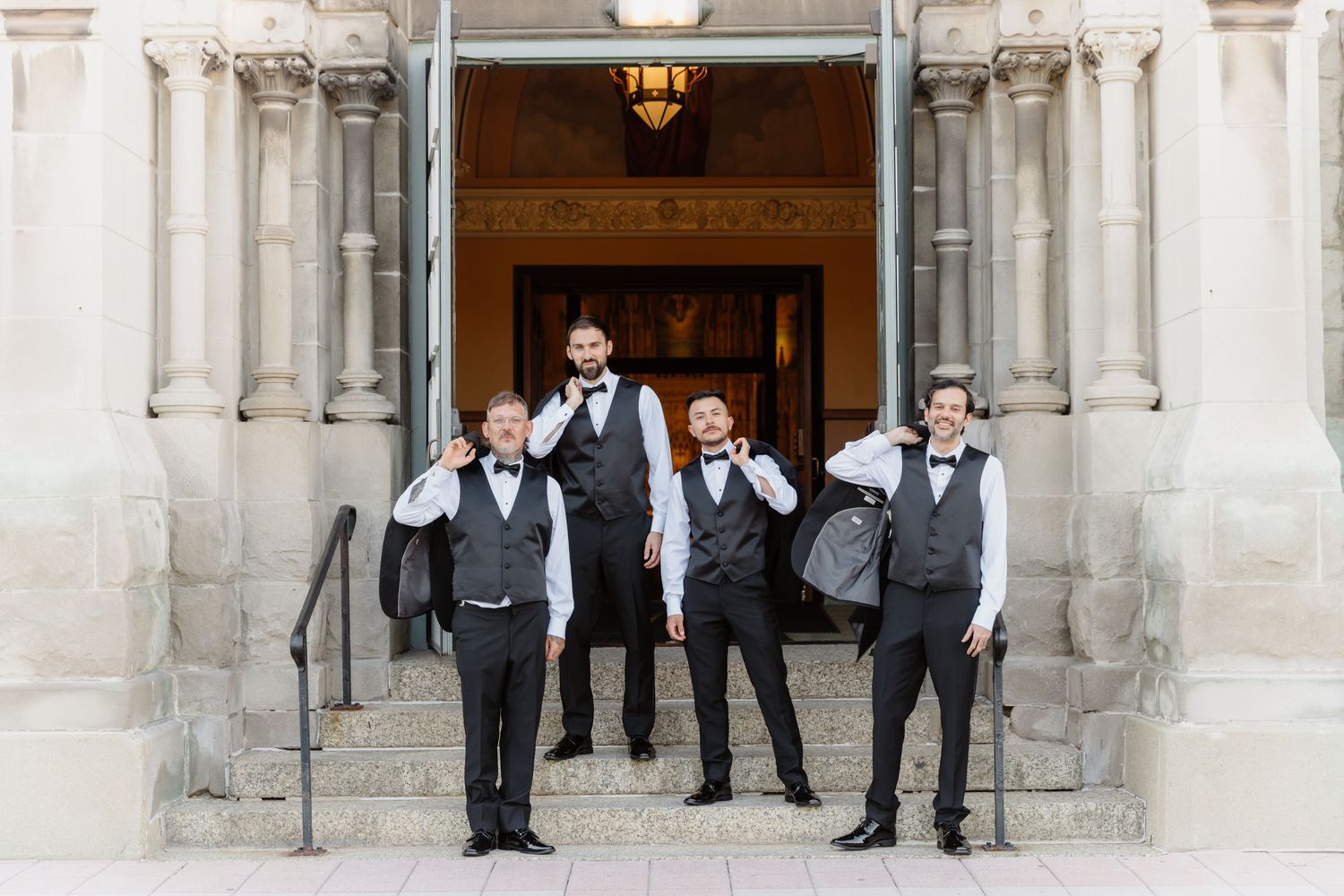 Four groomsmen pose playfully on stone steps outside an elegant building in matching black suits and bow ties.