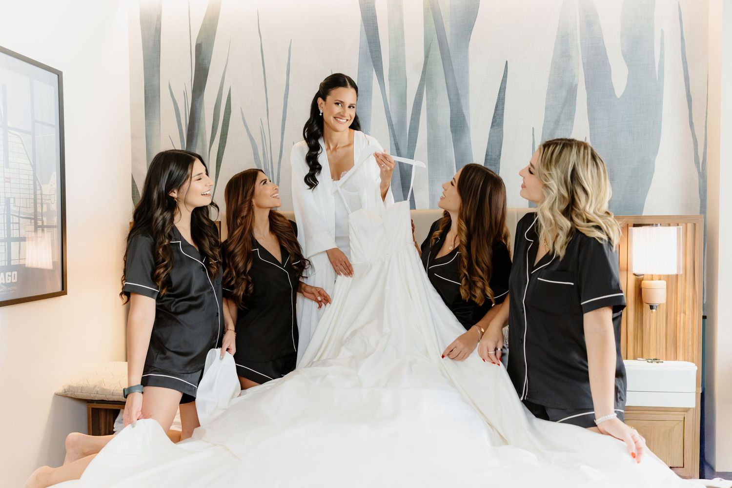 Bride in white robe holds gown while bridesmaids in black pajamas admire it on hotel bed.