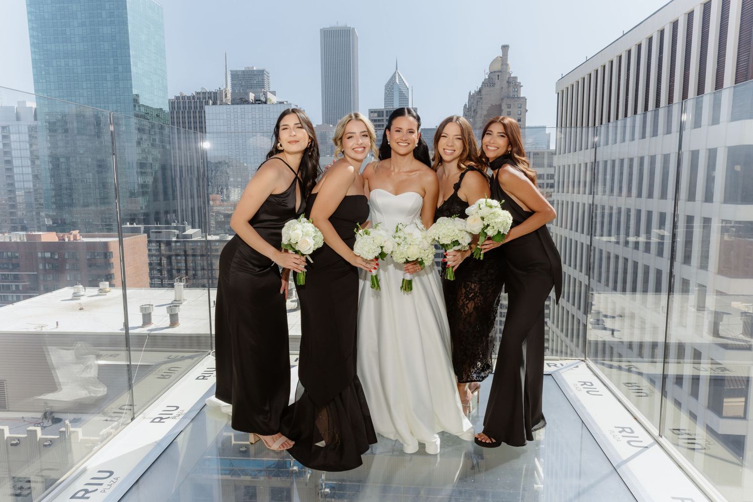 Smiling bride with bridesmaids in black gowns posing on rooftop at dusk in Chicago.