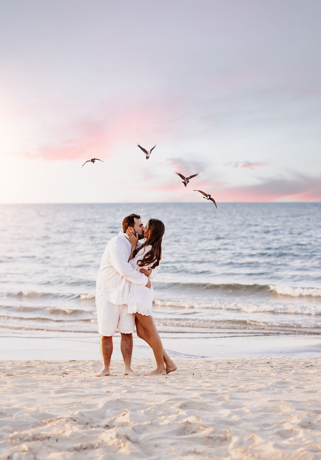 Couple kissing on beach at sunset with birds flying over calm ocean waves.