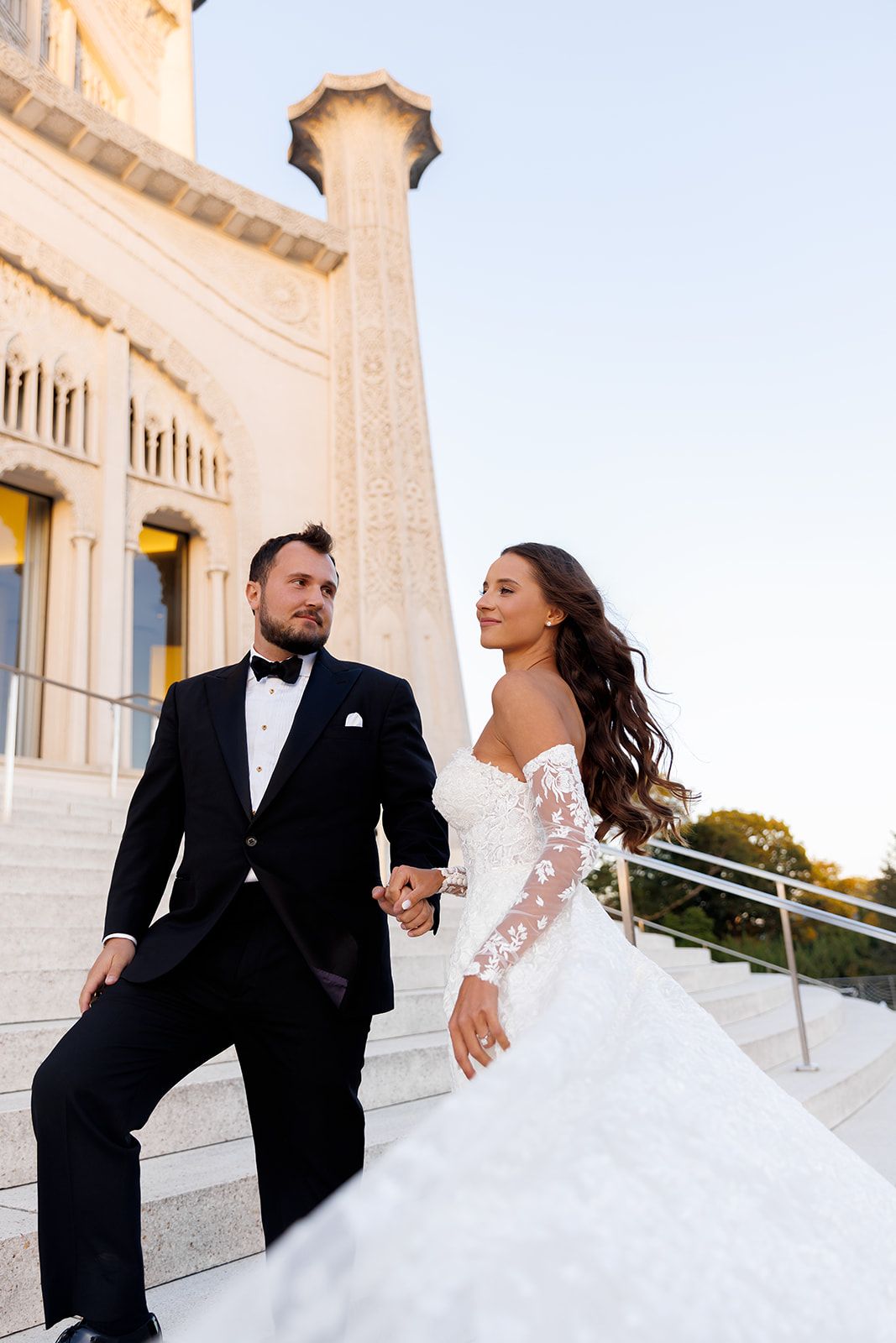 Bride and groom holding hands on grand stone steps of ornate building at sunset wedding.