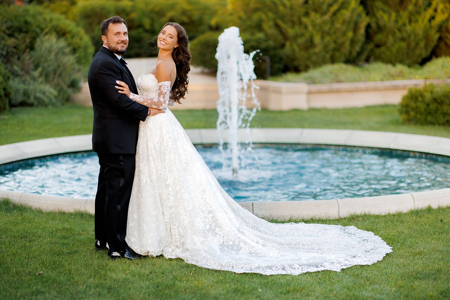 Bride and groom share romantic moment by garden fountain at dusk, bride in lace wedding dress.