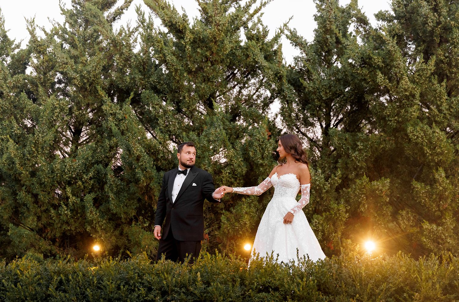 Bride and groom hold hands among tall evergreen trees with warm glowing lights at their wedding celebration.