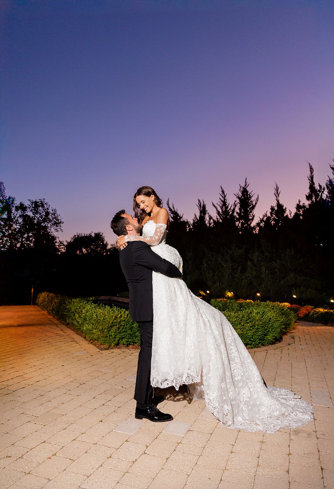 Groom lifts bride in elegant lace wedding gown at twilight, surrounded by manicured gardens under a purple sky.