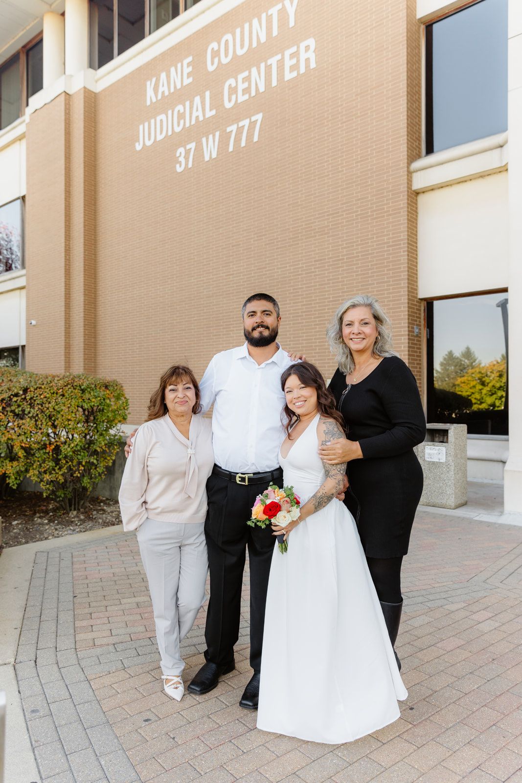 Wedding party of four poses outside Kane County Judicial Center, bride holding colorful bouquet.