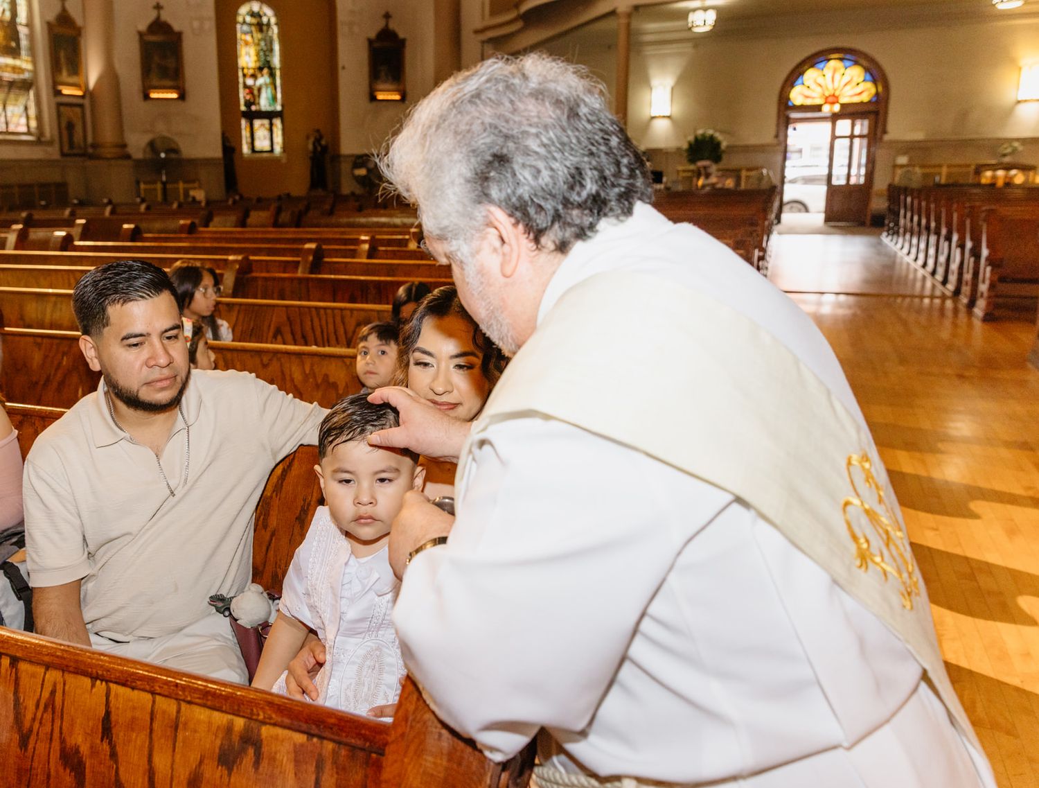 Catholic priest blesses young child with family during church ceremony in pew.