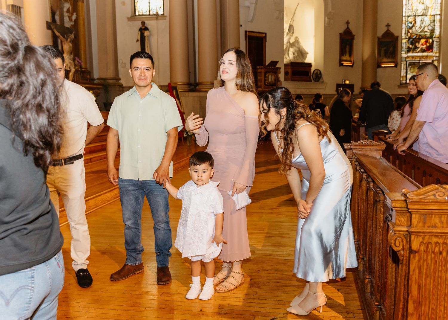Family gathered inside a church after a baptism, toddler in white dress standing between parents near wooden pews.